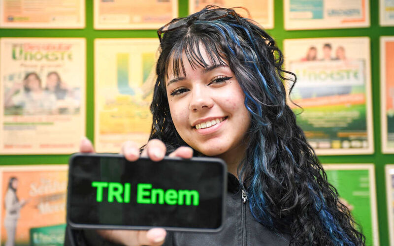 Estudante segura um celular com a frase “TRI Enem” exibida na tela, em frente a um painel de cartazes acadêmicos.