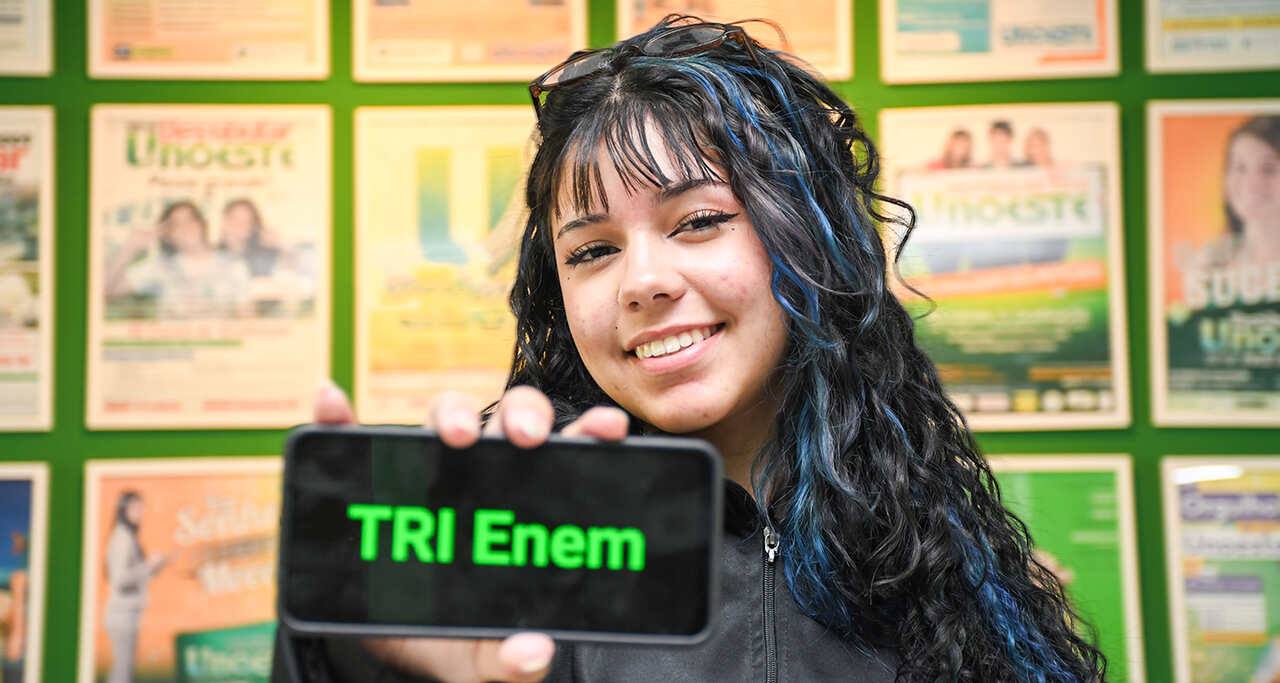 Estudante segura um celular com a frase “TRI Enem” exibida na tela, em frente a um painel de cartazes acadêmicos.