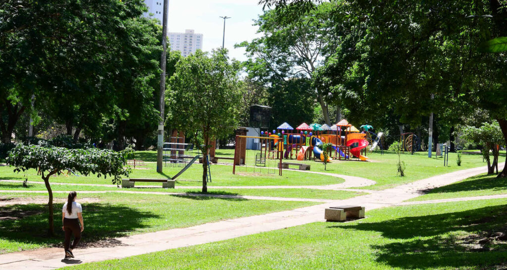Área verde do Parque do Povo, em Presidente Prudente, com caminhos pavimentados, árvores, playground colorido e pessoa caminhando pelo parque.