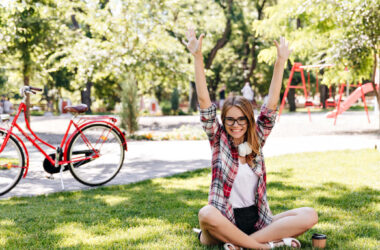 Jovem sentada no gramado de um parque arborizado, com os braços levantados, enquanto uma bicicleta vermelha está parada ao fundo perto de árvores e playground.