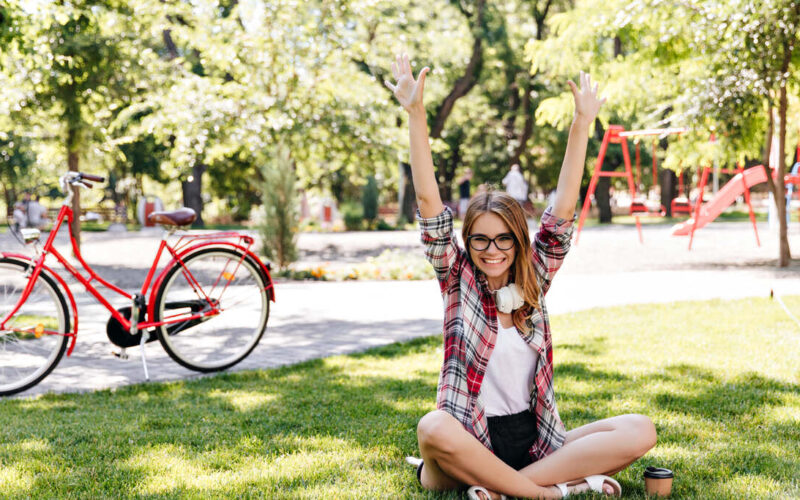 Jovem sentada no gramado de um parque arborizado, com os braços levantados, enquanto uma bicicleta vermelha está parada ao fundo perto de árvores e playground.