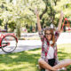 Jovem sentada no gramado de um parque arborizado, com os braços levantados, enquanto uma bicicleta vermelha está parada ao fundo perto de árvores e playground.