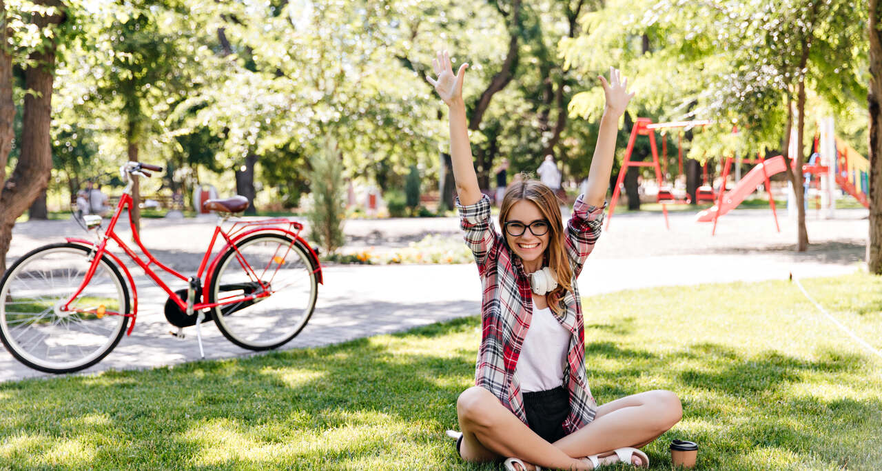 Jovem sentada no gramado de um parque arborizado, com os braços levantados, enquanto uma bicicleta vermelha está parada ao fundo perto de árvores e playground.