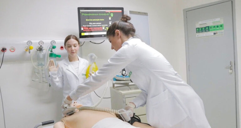 Alunas de Medicina da Unoeste realizam treinamento prático em um laboratório de simulação realística, utilizando manequim de alta fidelidade, desfibrilador e monitor cardíaco durante exercício de atendimento de emergência.