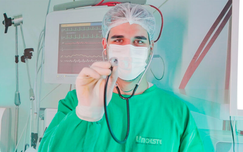 Estudante de Medicina com jaleco cirúrgico e estetoscópio em ambiente hospitalar, simbolizando a prática médica e o cuidado com a saúde.