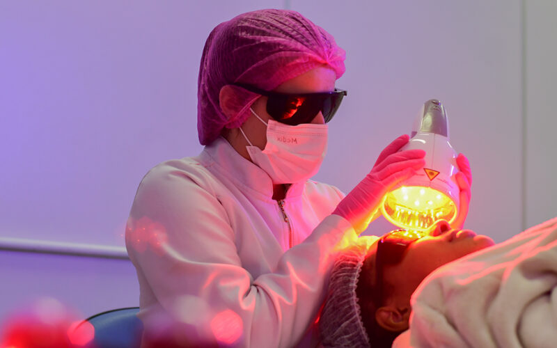 Aluna da faculdade de Estética e Cosmética realiza procedimento facial não invasivo em aula prática, utilizando equipamento estético e seguindo normas de biossegurança.