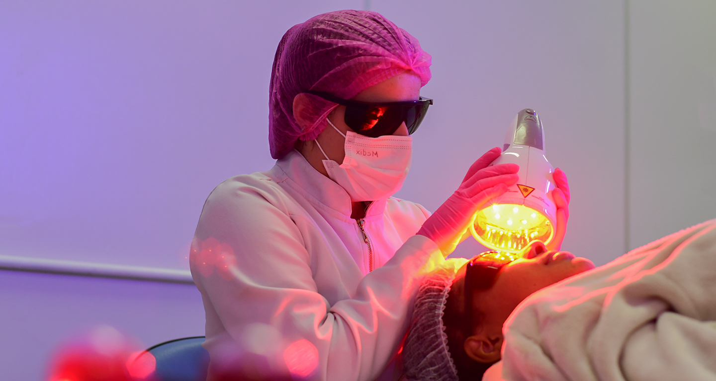 Aluna da faculdade de Estética e Cosmética realiza procedimento facial não invasivo em aula prática, utilizando equipamento estético e seguindo normas de biossegurança.