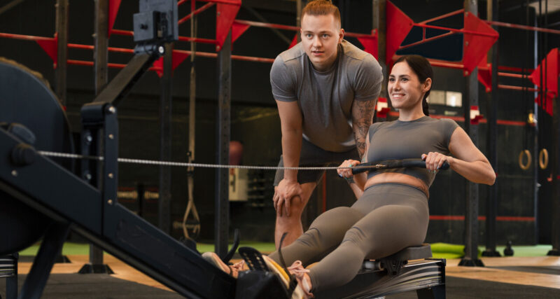 Profissional de Educação Física orientando aluna durante exercício em equipamento de treino funcional dentro de academia, com foco em saúde e desempenho físico.
