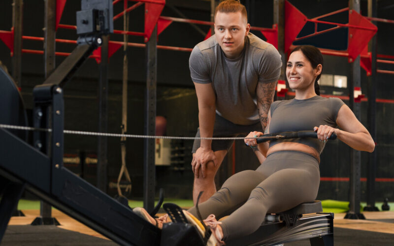 Profissional de Educação Física orientando aluna durante exercício em equipamento de treino funcional dentro de academia, com foco em saúde e desempenho físico.