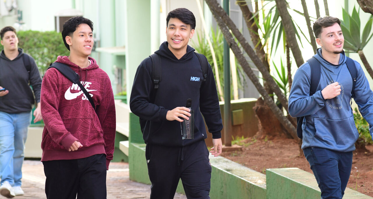 Três jovens utilizando mochilas e sorrindo caminham pelo campus da Unoeste.