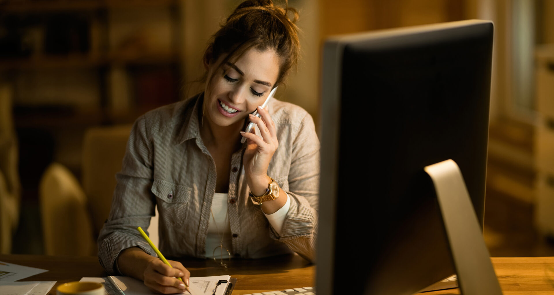 Mulher sentada à mesa, trabalhando em home office, falando ao telefone enquanto faz anotações em um caderno, com computador ligado à sua frente em ambiente interno.