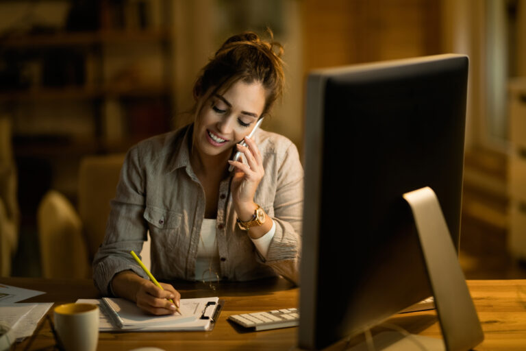 Mulher sentada à mesa, trabalhando em home office, falando ao telefone enquanto faz anotações em um caderno, com computador ligado à sua frente em ambiente interno.