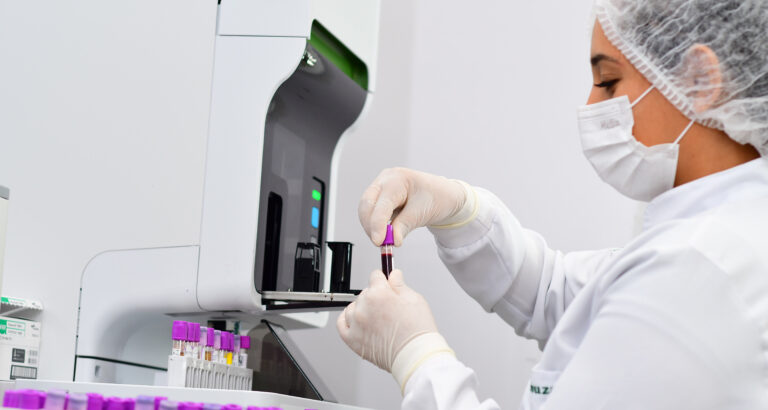 Profissional da saúde usando luvas, segurando tubo com amostra de sangue em ambiente de laboratório.
