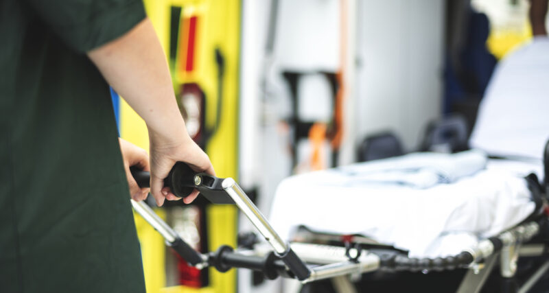 Profissional da área da saúde segurando a maca de uma ambulância, com equipamentos de atendimento pré-hospitalar visíveis ao fundo e interior do veículo desfocado.