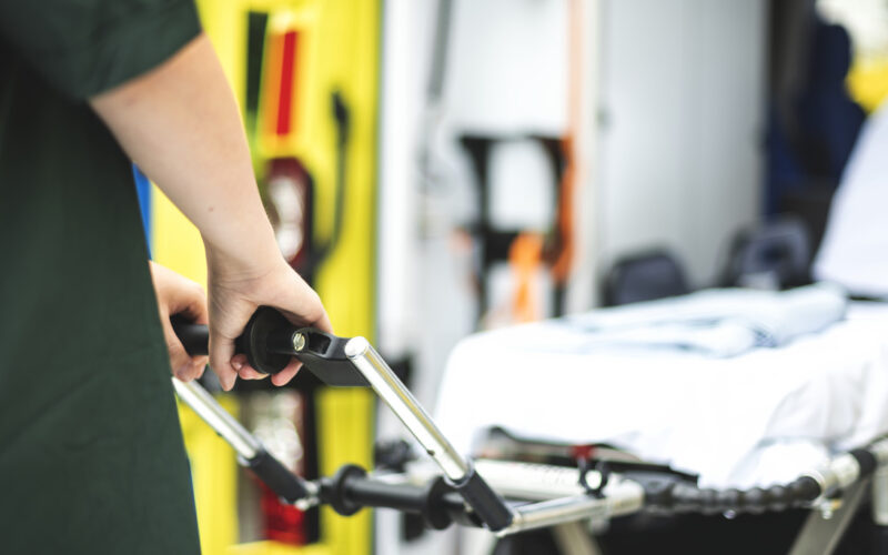 Profissional da área da saúde segurando a maca de uma ambulância, com equipamentos de atendimento pré-hospitalar visíveis ao fundo e interior do veículo desfocado.