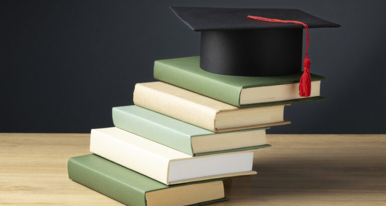 Pilha de livros sobre uma mesa, com um capelo de formatura preto e tassel vermelho no topo, simbolizando a jornada de conhecimento, a excelência acadêmica e a dedicação do corpo docente da Unoeste na formação de futuros profissionais.