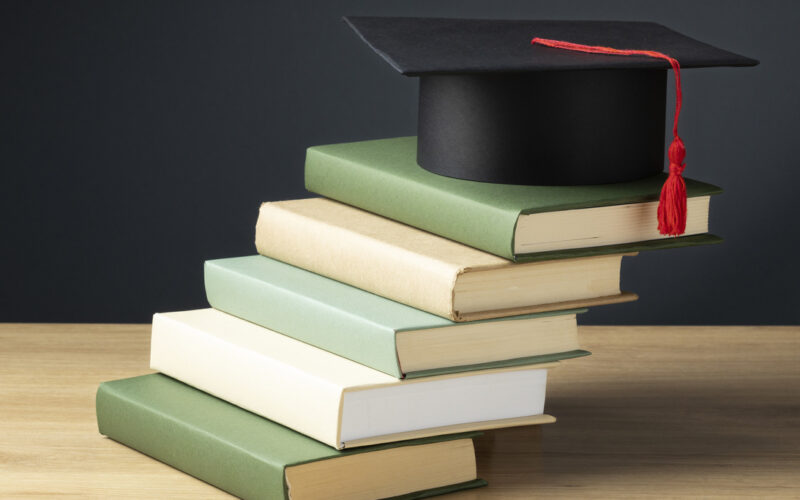 Pilha de livros sobre uma mesa, com um capelo de formatura preto e tassel vermelho no topo, simbolizando a jornada de conhecimento, a excelência acadêmica e a dedicação do corpo docente da Unoeste na formação de futuros profissionais.