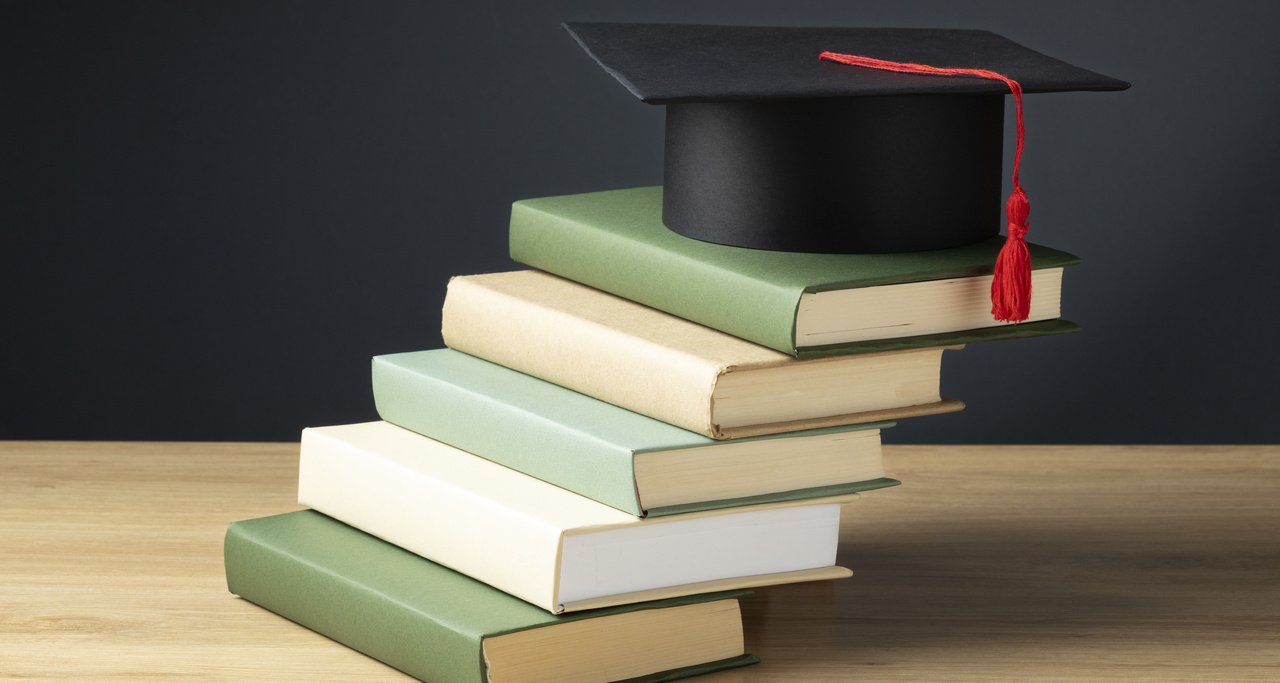 Pilha de livros sobre uma mesa, com um capelo de formatura preto e tassel vermelho no topo, simbolizando a jornada de conhecimento, a excelência acadêmica e a dedicação do corpo docente da Unoeste na formação de futuros profissionais.