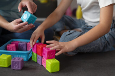 Criança sentada no chão organiza blocos coloridos enquanto um adulto segura um cubo educativo durante atividade lúdica em ambiente terapêutico.