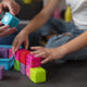 Criança sentada no chão organiza blocos coloridos enquanto um adulto segura um cubo educativo durante atividade lúdica em ambiente terapêutico.