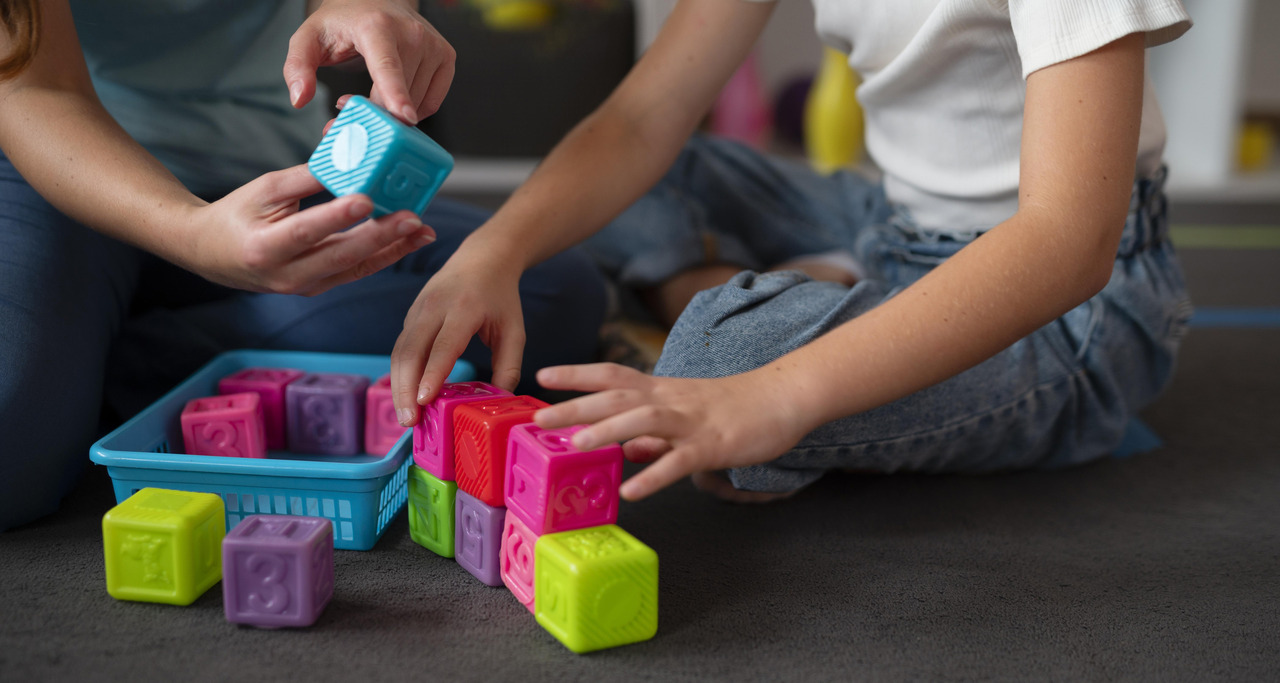 Criança sentada no chão organiza blocos coloridos enquanto um adulto segura um cubo educativo durante atividade lúdica em ambiente terapêutico.