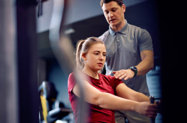 Personal trainer acompanha jovem durante exercício em máquina de musculação na academia, orientando postura com a mão no ombro enquanto ela realiza o movimento sentada, em ambiente interno com equipamentos ao fundo.