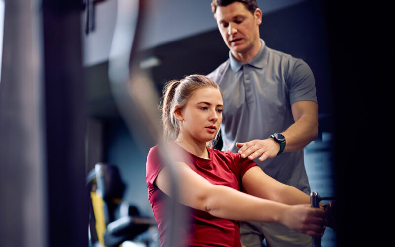 Personal trainer acompanha jovem durante exercício em máquina de musculação na academia, orientando postura com a mão no ombro enquanto ela realiza o movimento sentada, em ambiente interno com equipamentos ao fundo.