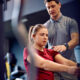 Personal trainer acompanha jovem durante exercício em máquina de musculação na academia, orientando postura com a mão no ombro enquanto ela realiza o movimento sentada, em ambiente interno com equipamentos ao fundo.