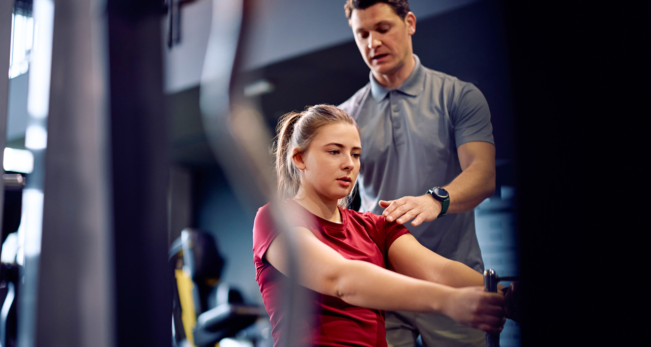 Personal trainer acompanha jovem durante exercício em máquina de musculação na academia, orientando postura com a mão no ombro enquanto ela realiza o movimento sentada, em ambiente interno com equipamentos ao fundo.