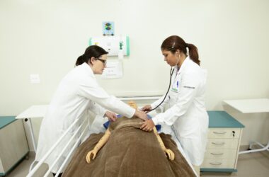 Estudantes de Enfermagem em aula prática realizam avaliação clínica em manequim hospitalar, utilizando estetoscópio em laboratório de simulação.