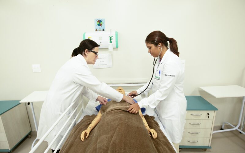 Estudantes de Enfermagem em aula prática realizam avaliação clínica em manequim hospitalar, utilizando estetoscópio em laboratório de simulação.