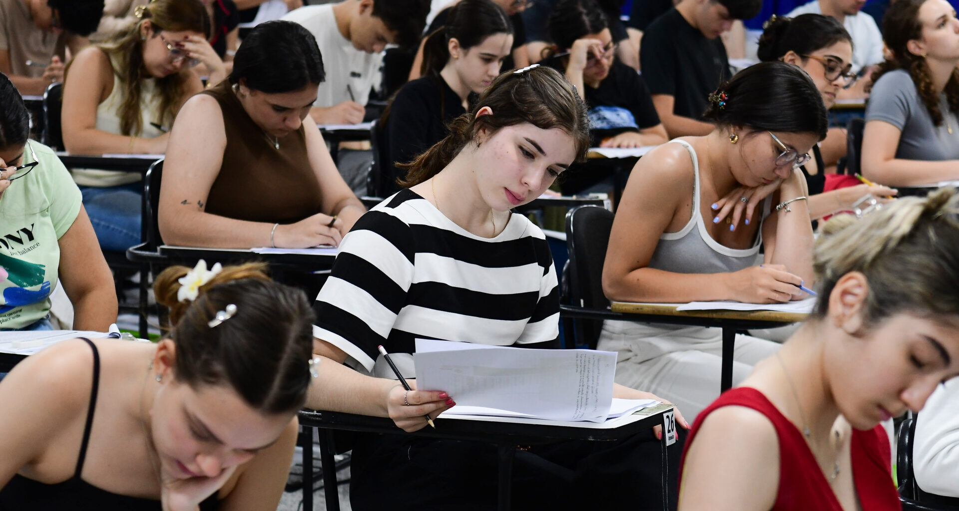 Estudantes concentrados realizando prova de vestibular em sala de aula, escrevendo e analisando questões nas folhas, demonstrando foco, gestão de tempo e preparação para o exame.