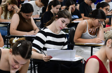 Estudantes concentrados realizando prova de vestibular em sala de aula, escrevendo e analisando questões nas folhas, demonstrando foco, gestão de tempo e preparação para o exame.