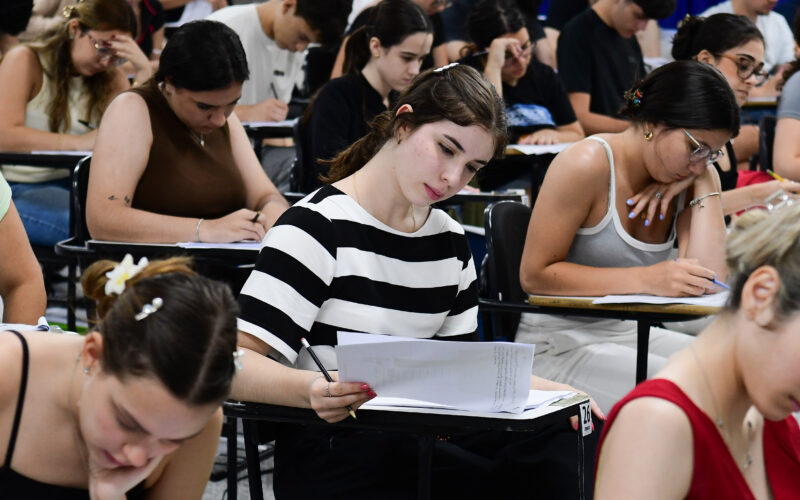 Estudantes concentrados realizando prova de vestibular em sala de aula, escrevendo e analisando questões nas folhas, demonstrando foco, gestão de tempo e preparação para o exame.
