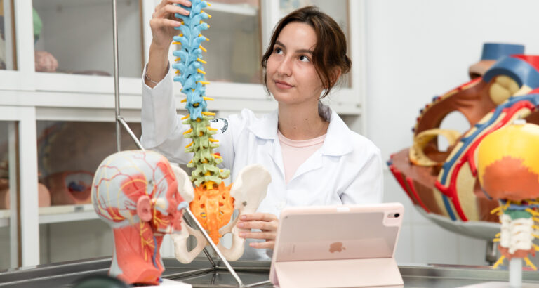 Estudante da área da saúde observa modelo anatômico da coluna vertebral e pelve em laboratório, ilustrando o ambiente de aprendizado do curso de Medicina.