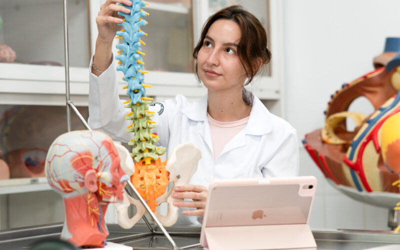 Estudante da área da saúde observa modelo anatômico da coluna vertebral e pelve em laboratório, ilustrando o ambiente de aprendizado do curso de Medicina.