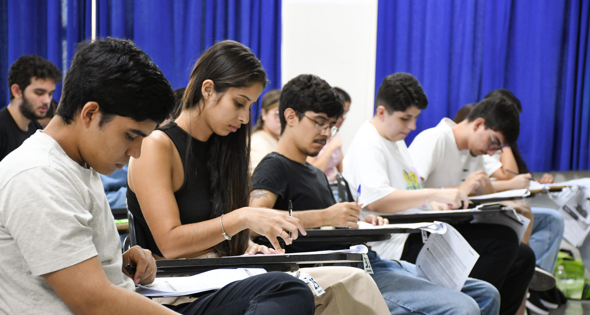 Estudantes sentados em fileiras em uma sala de aula durante a realização de uma prova de vestibular, concentrados enquanto escrevem nas folhas de resposta apoiadas nas carteiras, com cortinas azuis ao fundo.
