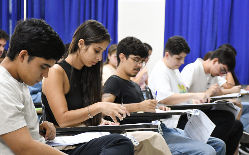 Estudantes sentados em fileiras em uma sala de aula durante a realização de uma prova de vestibular, concentrados enquanto escrevem nas folhas de resposta apoiadas nas carteiras, com cortinas azuis ao fundo.