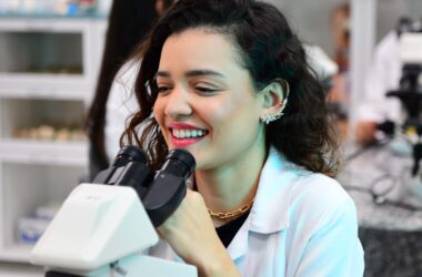 Estudante de Biomedicina da Unoeste em laboratório utilizando microscópio, sorrindo durante aula prática, com outros alunos e equipamentos científicos ao fundo.