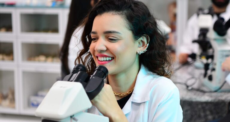 Estudante de Biomedicina da Unoeste em laboratório utilizando microscópio, sorrindo durante aula prática, com outros alunos e equipamentos científicos ao fundo.