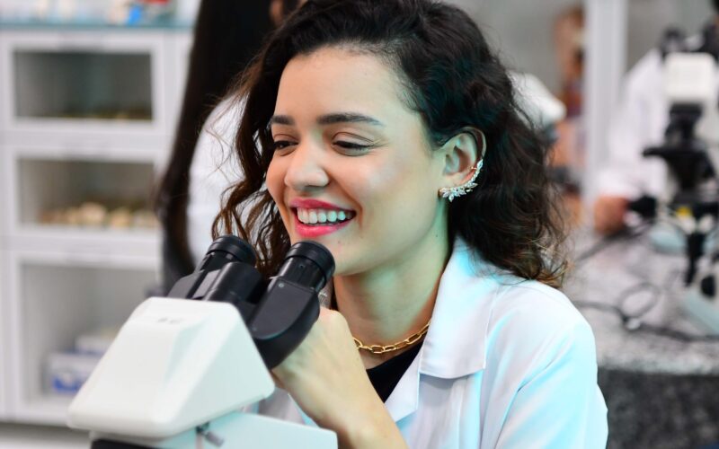 Estudante de Biomedicina da Unoeste em laboratório utilizando microscópio, sorrindo durante aula prática, com outros alunos e equipamentos científicos ao fundo.