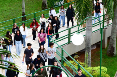 Grupo de estudantes caminhando por uma área externa de campus universitário, em uma passagem com rampas e corrimãos verdes, cercada por árvores e gramado, enquanto conversam e seguem juntos para atividades acadêmicas.