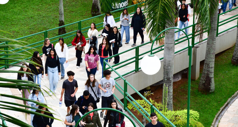 Grupo de estudantes caminhando por uma área externa de campus universitário, em uma passagem com rampas e corrimãos verdes, cercada por árvores e gramado, enquanto conversam e seguem juntos para atividades acadêmicas.
