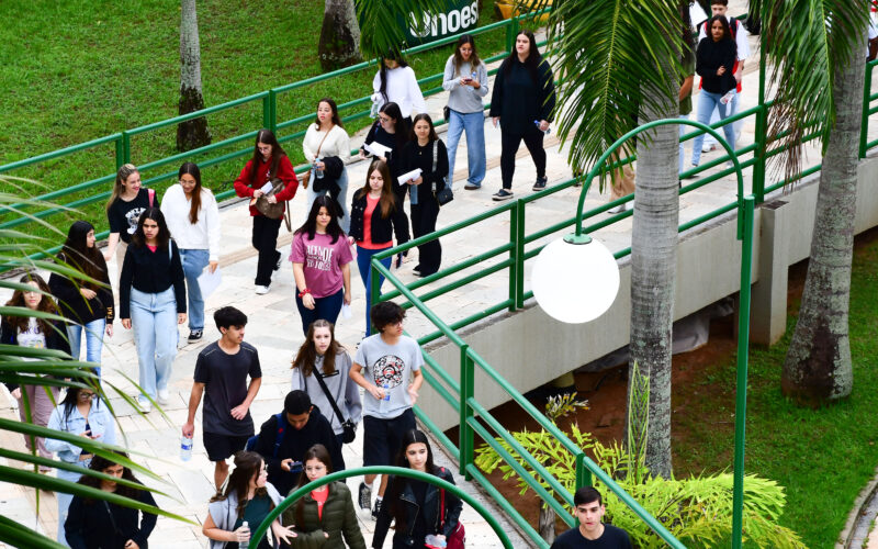 Grupo de estudantes caminhando por uma área externa de campus universitário, em uma passagem com rampas e corrimãos verdes, cercada por árvores e gramado, enquanto conversam e seguem juntos para atividades acadêmicas.