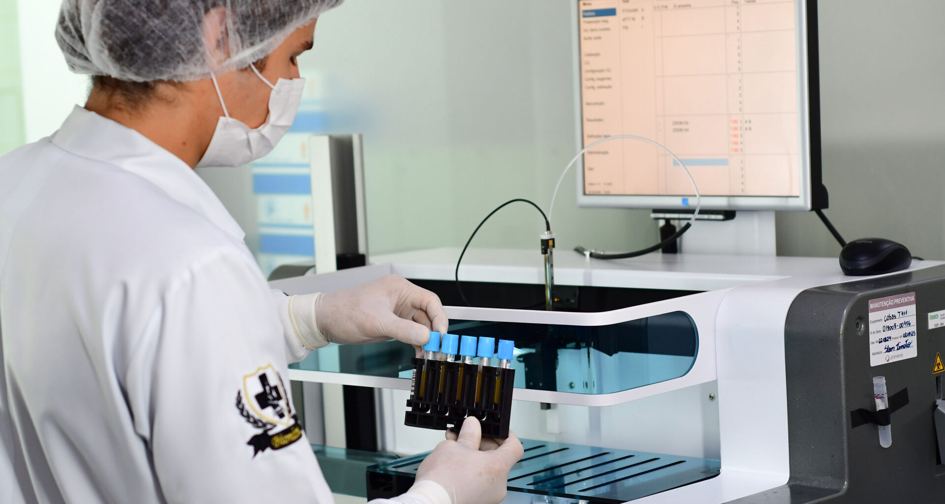 Profissional em laboratório utiliza equipamentos automatizados para análise de amostras, segurando tubos de ensaio com luvas, máscara e touca, em ambiente controlado com monitor exibindo dados laboratoriais.