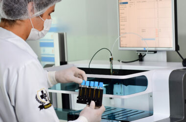 Profissional em laboratório utiliza equipamentos automatizados para análise de amostras, segurando tubos de ensaio com luvas, máscara e touca, em ambiente controlado com monitor exibindo dados laboratoriais.