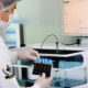 Profissional em laboratório utiliza equipamentos automatizados para análise de amostras, segurando tubos de ensaio com luvas, máscara e touca, em ambiente controlado com monitor exibindo dados laboratoriais.