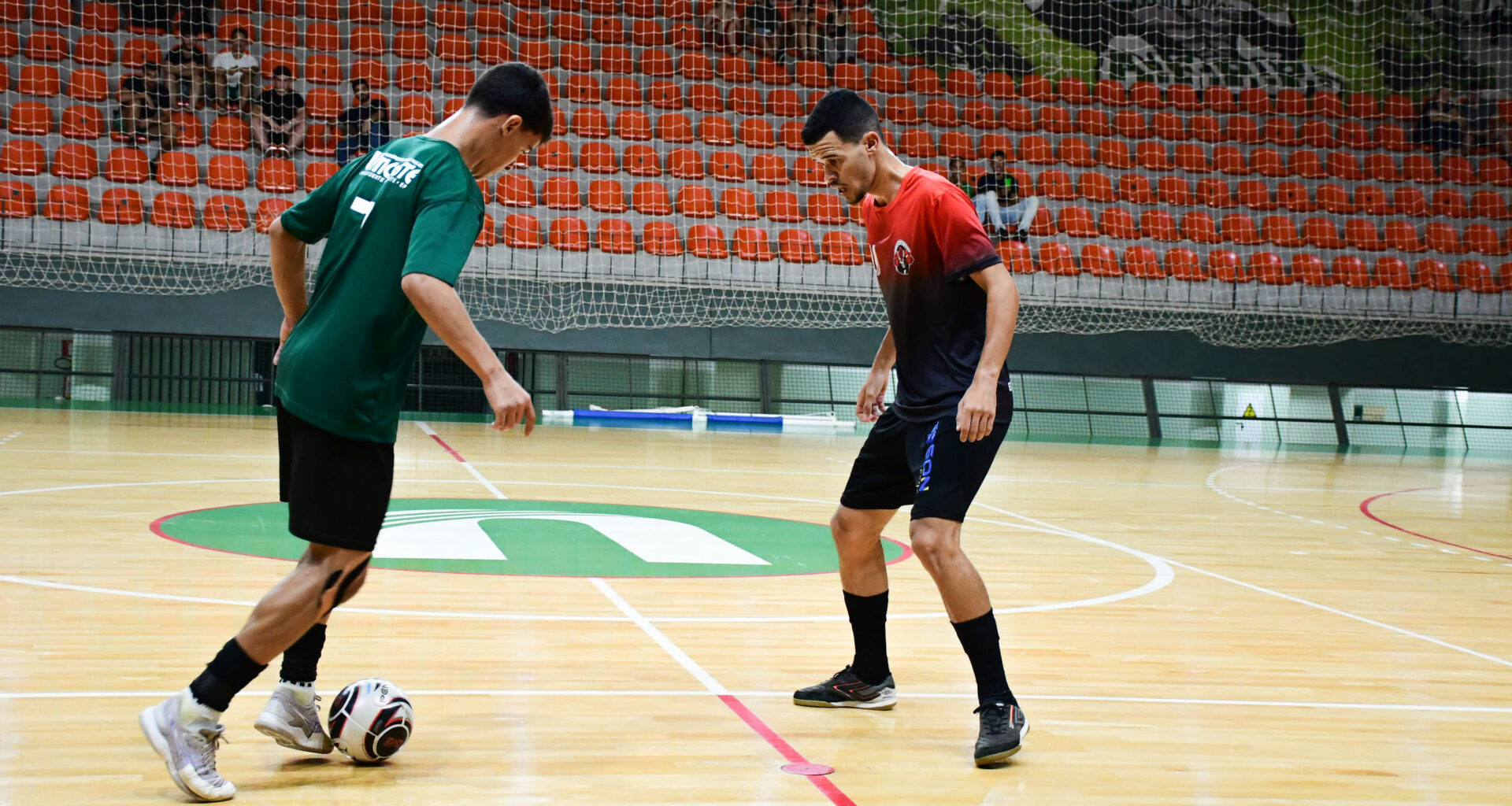 Dois jovens atletas jogam futsal em quadra coberta, disputando a bola com os pés em uma jogada próxima, enquanto mantêm postura de concentração, equilíbrio corporal e atenção aos movimentos do adversário durante a partida.