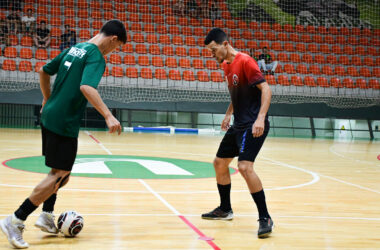 Dois jovens atletas jogam futsal em quadra coberta, disputando a bola com os pés em uma jogada próxima, enquanto mantêm postura de concentração, equilíbrio corporal e atenção aos movimentos do adversário durante a partida.