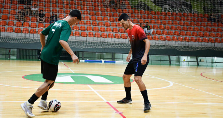 Dois jovens atletas jogam futsal em quadra coberta, disputando a bola com os pés em uma jogada próxima, enquanto mantêm postura de concentração, equilíbrio corporal e atenção aos movimentos do adversário durante a partida.